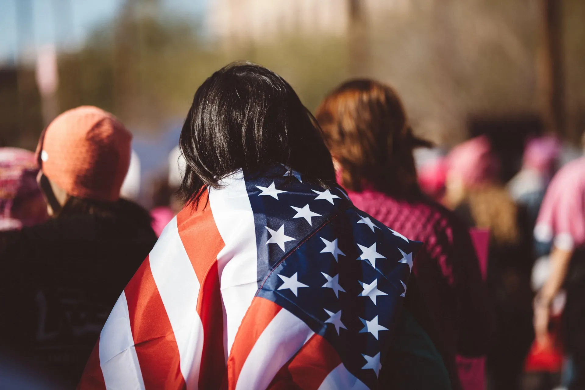 A crowd of people outdoors with only their backs visible and the person in the foreground wears an American flag draped around their shoulders.