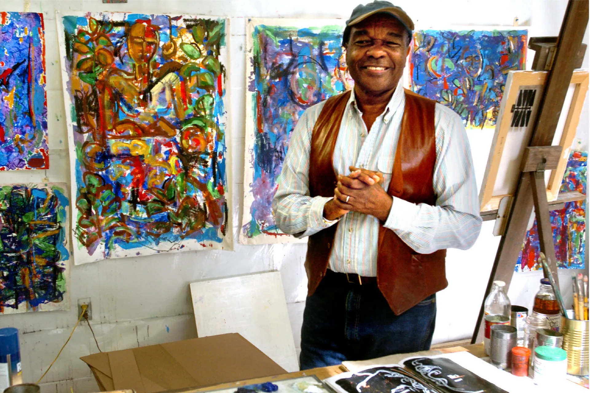 Artist David C. Driskell stands with hands clasped, smiling, in front of vibrant paintings displaying an array of color and shapes.